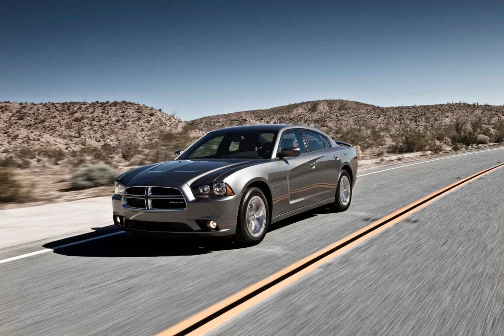 A dark grey metallic 2012 Dodge Charger drives on a paved road lined with desert brush and small hills