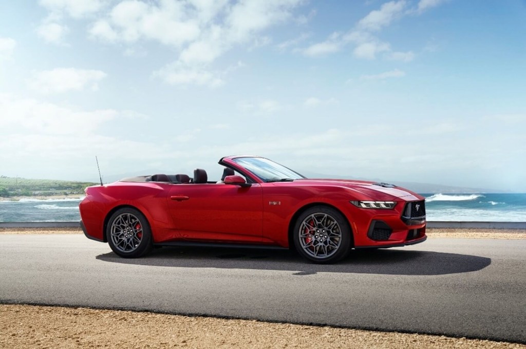 A red 2024 Ford Mustang Convertible, a GT drop-top with adaptive cruise control, parks next to a coastline. 