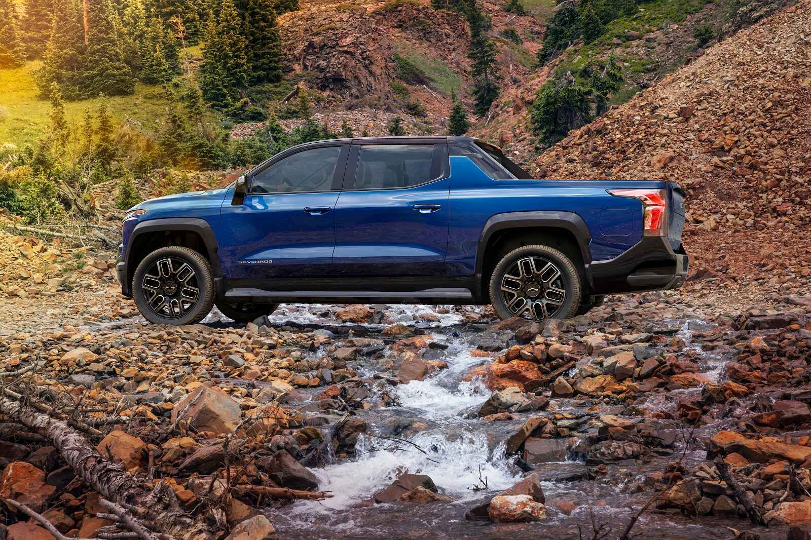 A blue 2024 Chevrolet Silverado EV parked in left profile view over a rocky creek