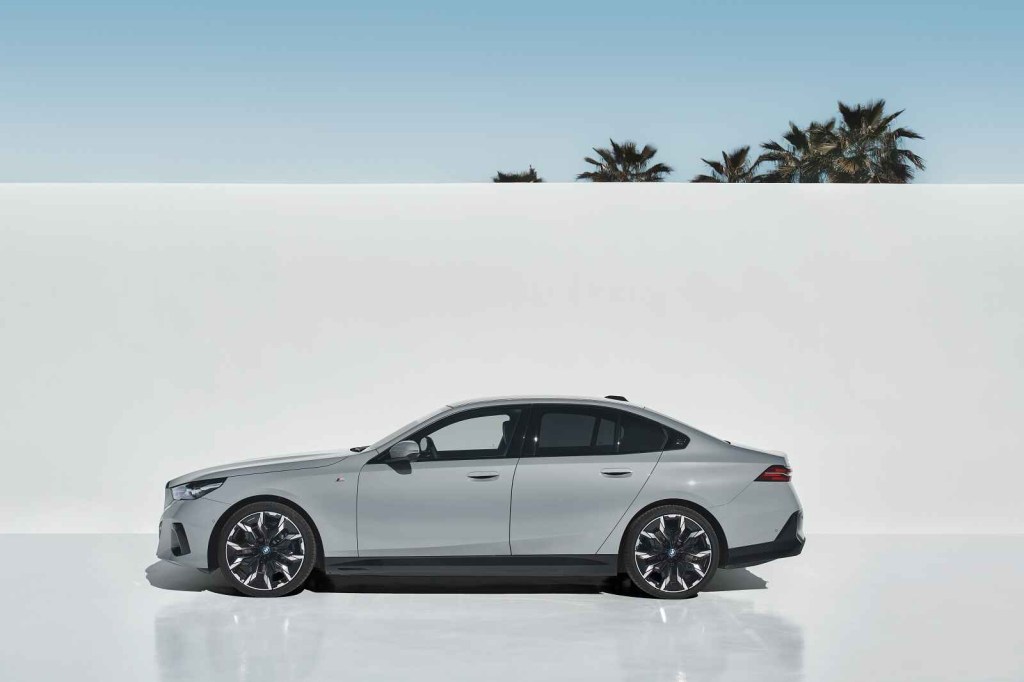 A silver 2024 BMW i5 EV sedan featured in a 2024 Super Bowl car commercial shown in full profile view with a white backdrop and palm trees peeking from top of backdrop