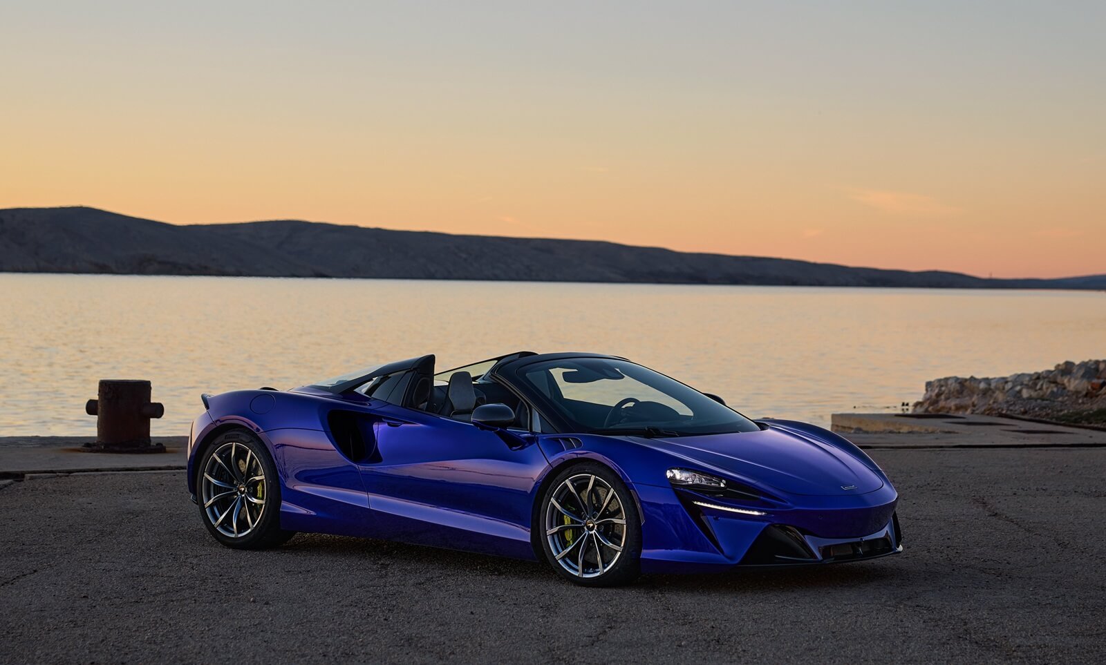 A blue 2025 McLaren Artura Spider parks next to the water. 