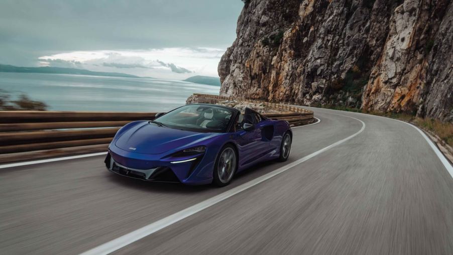 A blue 2025 McLaren Artura Spider cruises around a coastal road.