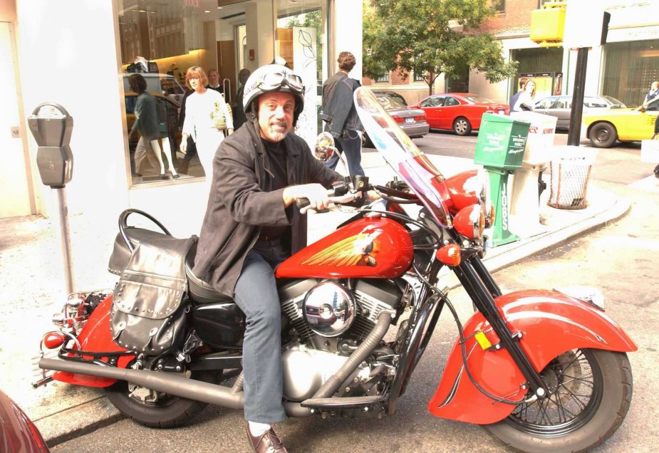 Musician Billy Joel sits on a red motorcycle, NYC visible in the background.