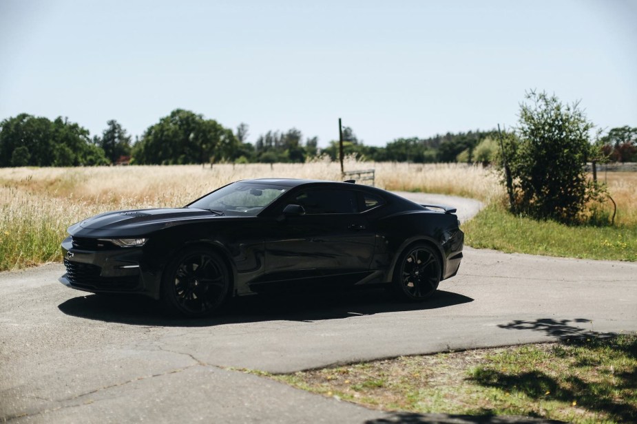 Black Camaro coupe parked on a paved road in front of an empty field and. trees.