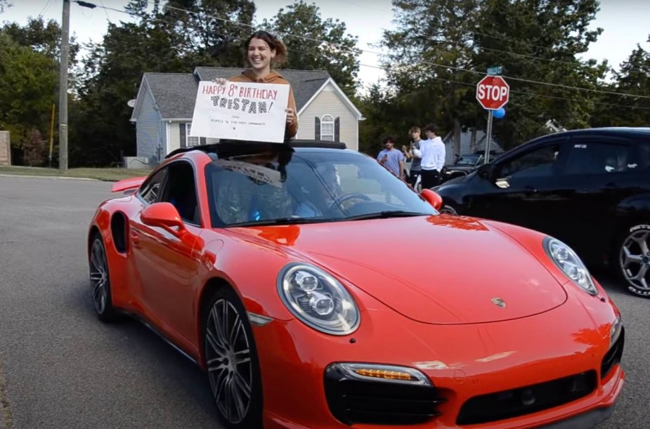 A woman holds a "happy birthday" sign atop a red Porsche during a parade.