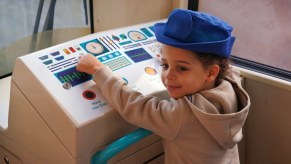 A little girl in a train driver hat sits at the pretend control console of a London commuter train.