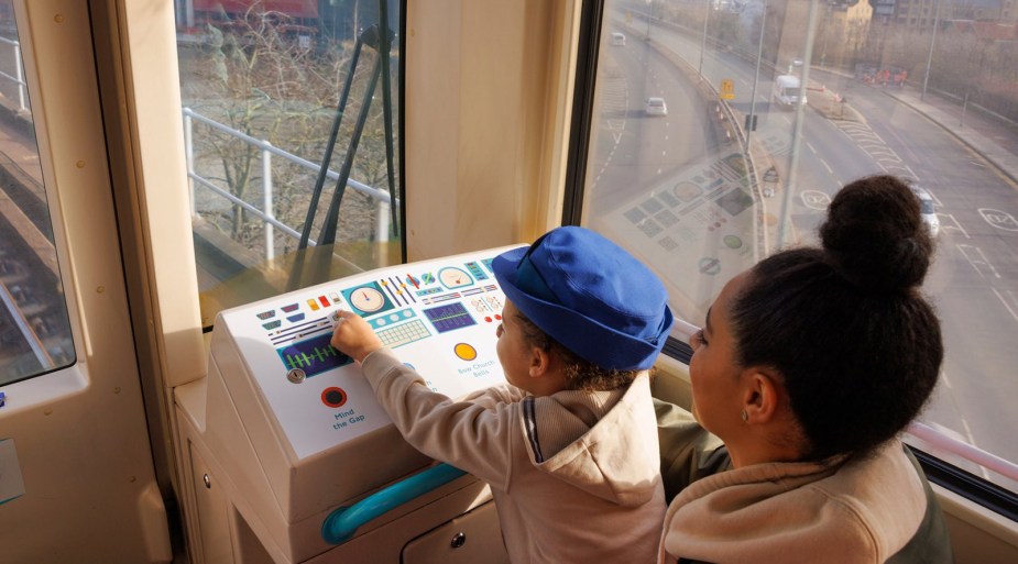 A little girl and her mother sit at the pretend driver console of a self-driving London train.