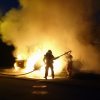 Firefighters battle a car fire with a vehicle like a Tesla or BMW.
