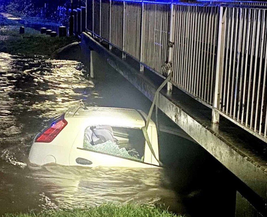 White compact car submerged in a flooded river, attached to a bridge with ratchet straps.