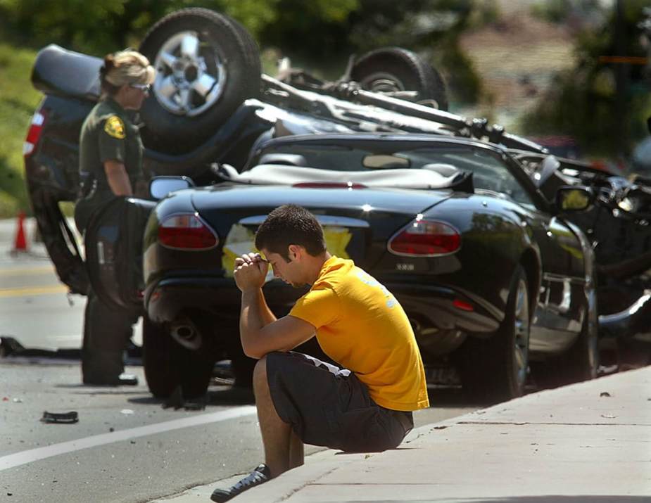 A motorist sits next to a Jaguar X100 XK and a totaled car after a crash.