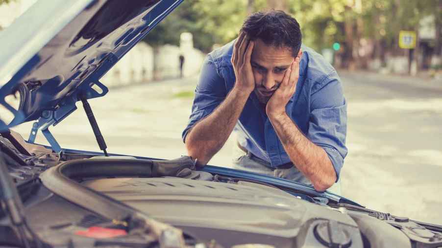 A man in a blue shirt visibly frustrated leaning over his open engine compartment