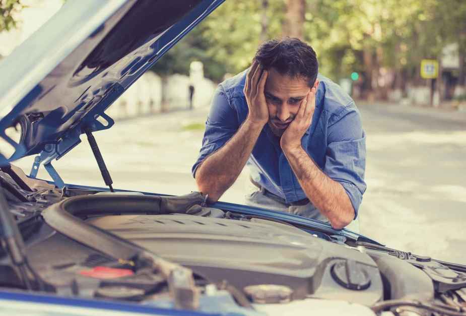A man in a blue shirt visibly frustrated leaning over his open engine compartment