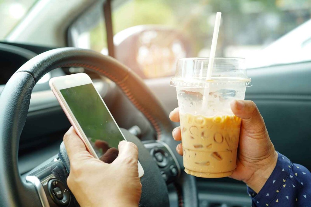 A man's hands hold an iced coffee drink and phone in front of car steering wheel close view