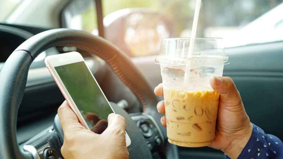 A man's hands hold an iced coffee drink and phone in front of car steering wheel close view