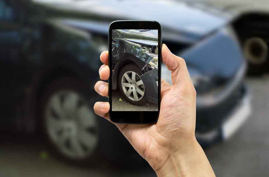 A hand holds up a cell phone taking a photo of a black car with right fender damage