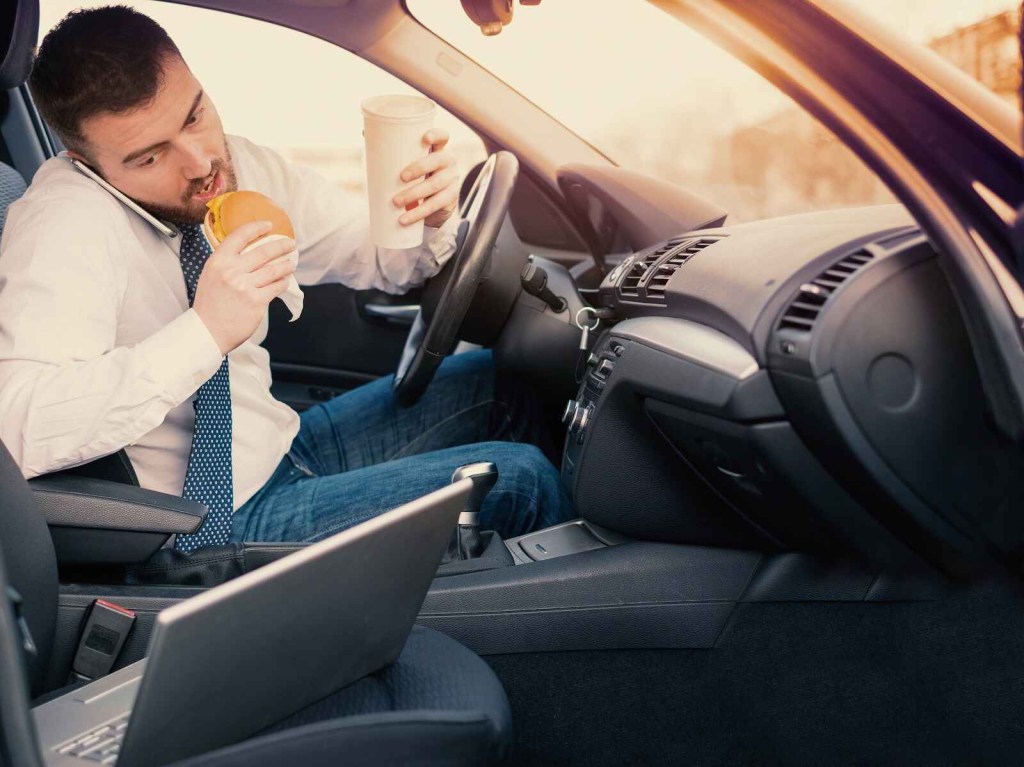 A man wearing a tie and jeans bites into a cheeseburger while cradling a cell phone on his neck and holding a drink in the driver's seat of a car