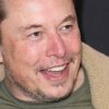Tesla CEO Elon Musk shown smiling wide in close angle view Elon looking to his left slightly wearing a shearling-lined jacket