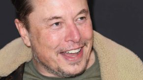 Tesla CEO Elon Musk shown smiling wide in close angle view Elon looking to his left slightly wearing a shearling-lined jacket