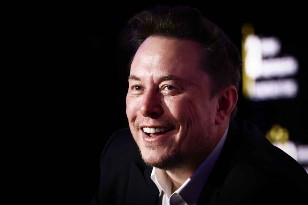 Tesla CEO Elon Musk sits in right profile angle view wearing a white collared shirt and dark jacket smiling