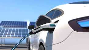 A white EV being charged with solar panels in the background in left rear view
