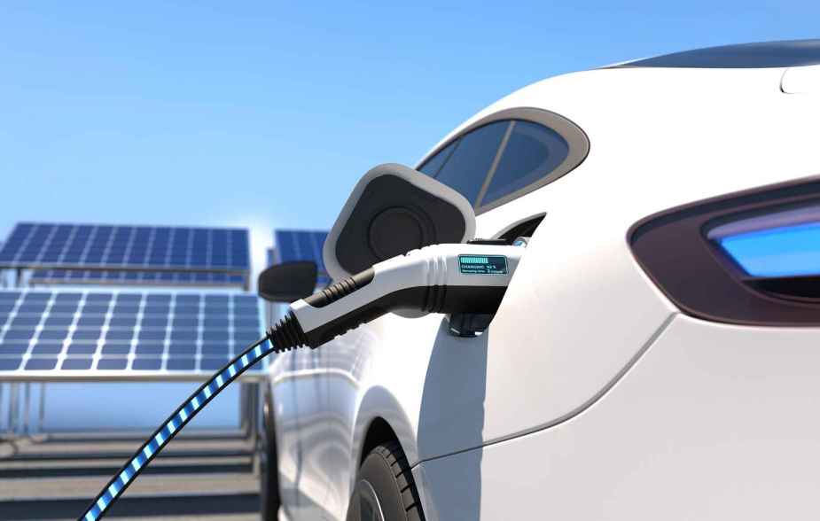 A white EV being charged with solar panels in the background in left rear view