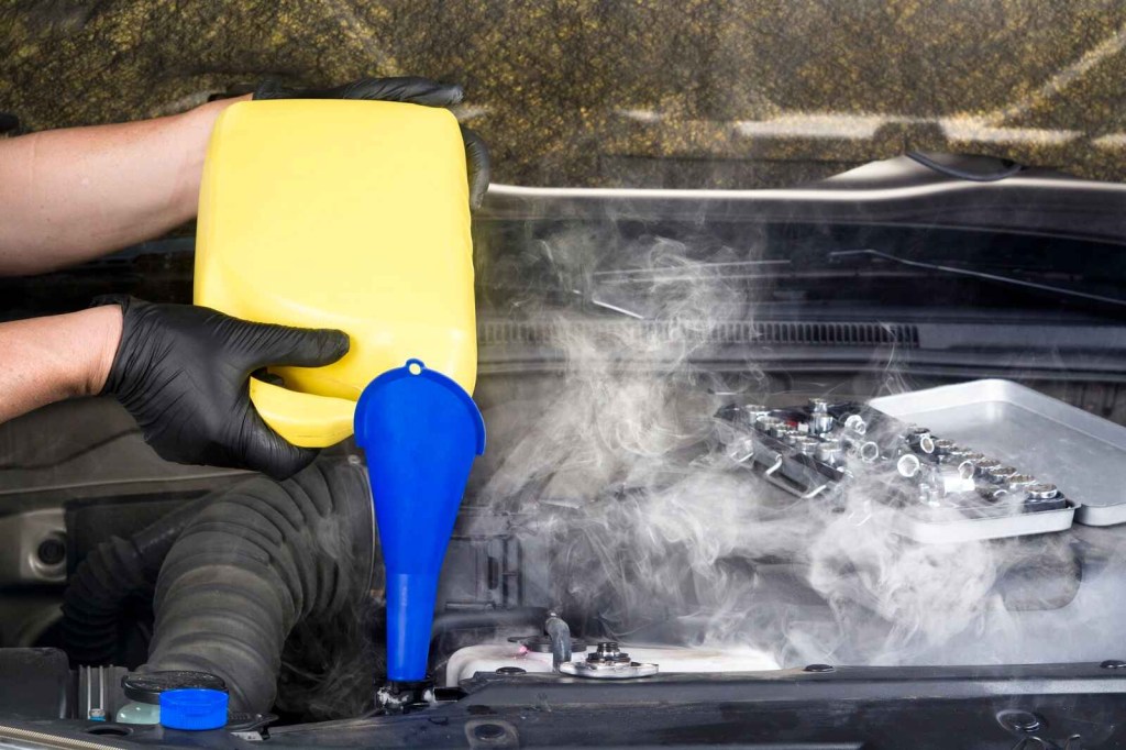 A gloved hand holds a container and pours it into a blue funnel into a steaming car radiator in close view