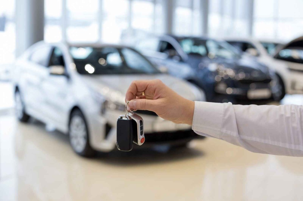 A man's hand holds up a car key in front of cars displayed in a showroom