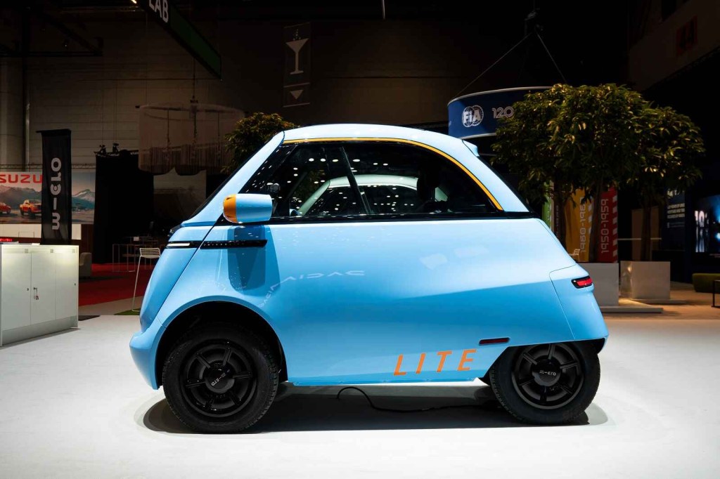 A blue Microlino Lite neighborhood electric bubble car is shown parked in full left profile view at the 2024 Geneva Auto Show