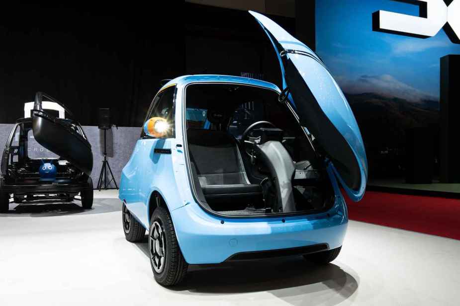 A blue Micro Microlino Lite NEV car with Isetta-like open front door is parked on display