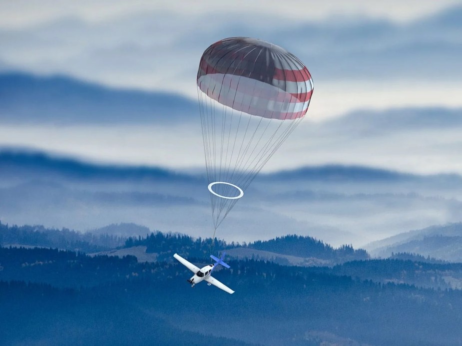 An ultralight Cirrus aircraft descending by parachute, mountains visible in the background.
