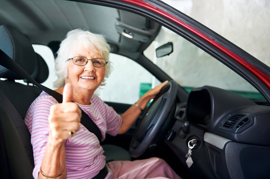An older woman gives a thumbs-up signal from the driver's seat of her car.