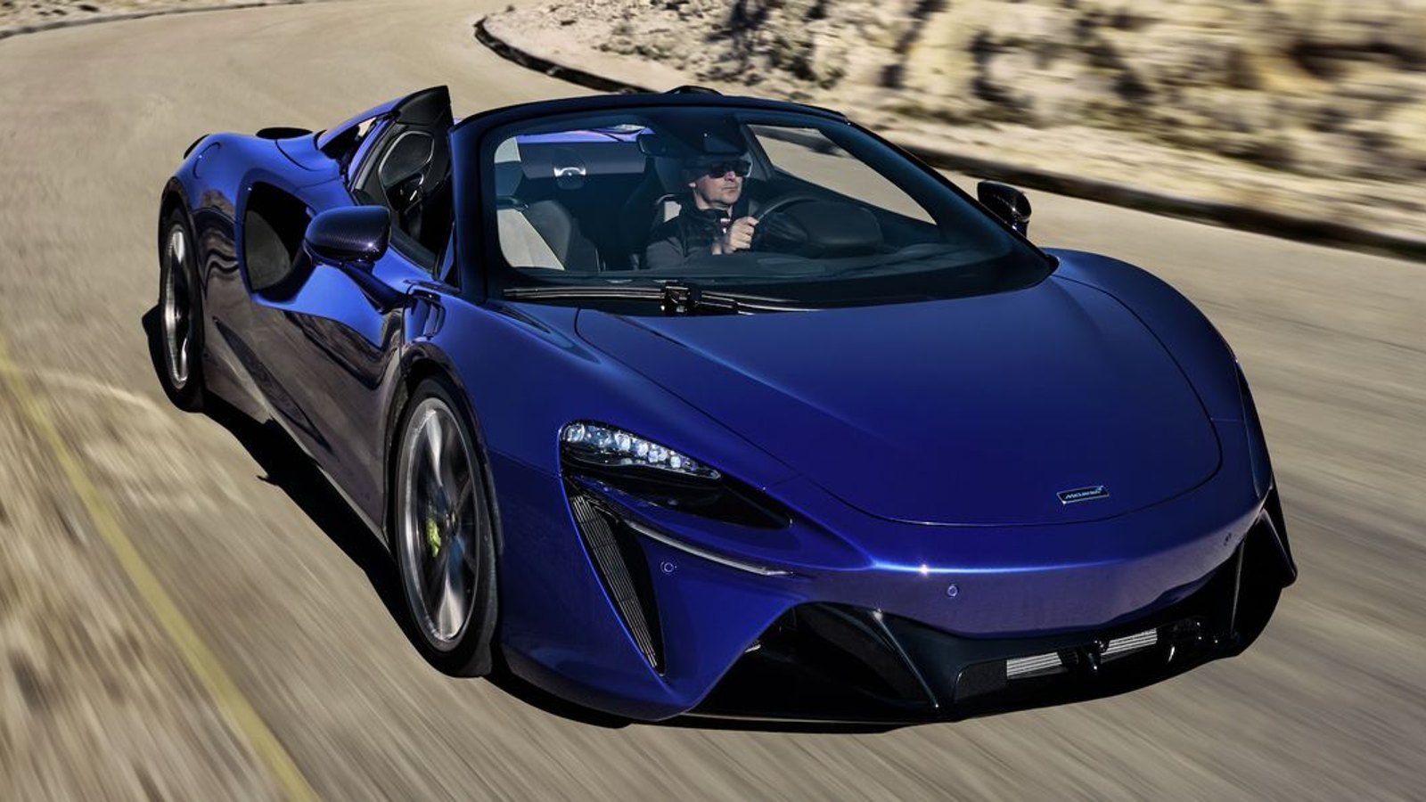 Blue McLaren Artura Spider supecar driving down a country road.