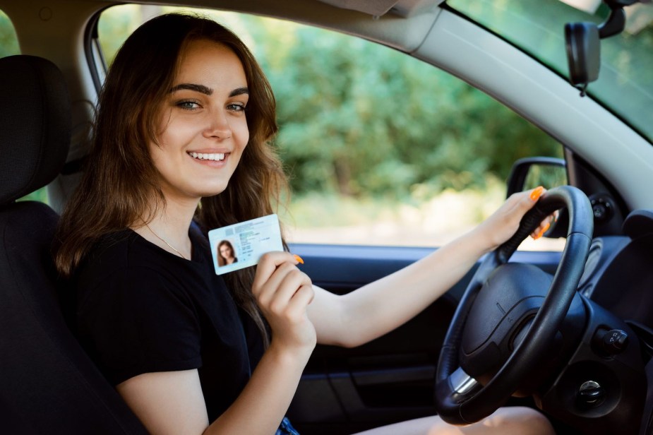 A young woman in the driver's seat of a car holds up her new license.