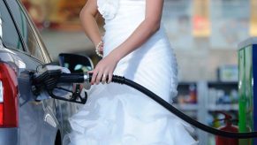 Woman from shoulders down wearing a white wedding dress shown refueling a car at a gas station left side view