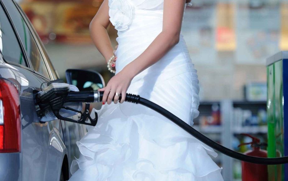 Woman from shoulders down wearing a white wedding dress shown refueling a car at a gas station left side view