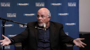 Finance "guru" Dave Ramsey sits with a shocked look on his face arms wide sitting in front of a studio microphone black suit jacket