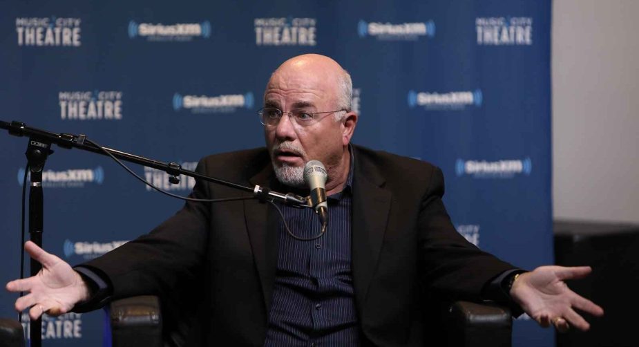 Finance "guru" Dave Ramsey sits with a shocked look on his face arms wide sitting in front of a studio microphone black suit jacket