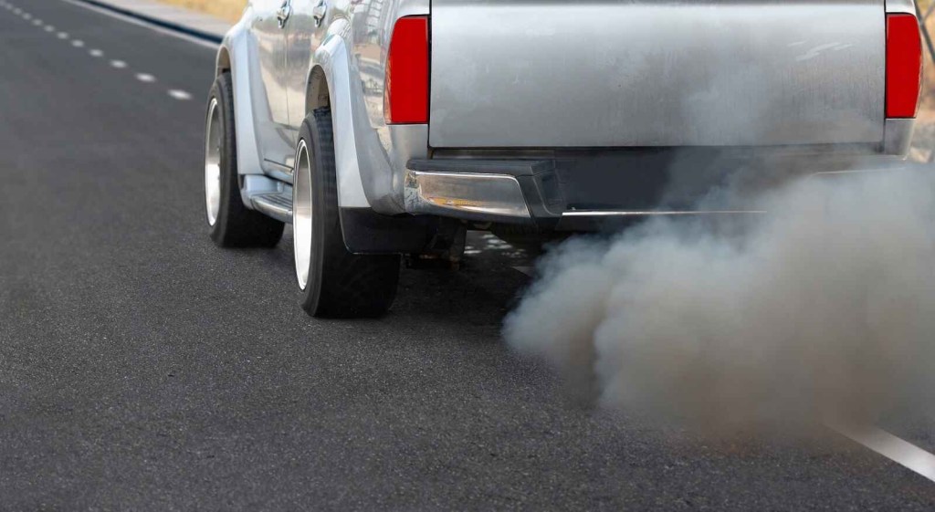 A silver diesel pickup truck driving on a paved road in close left rear angle blowing black exhaust smoke