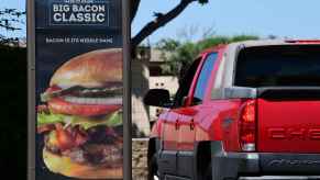 A red Chevrolet Avalanche truck stopped in a Wendy's drive-thru lane next to a large marketing sign for a bacon cheeseburger