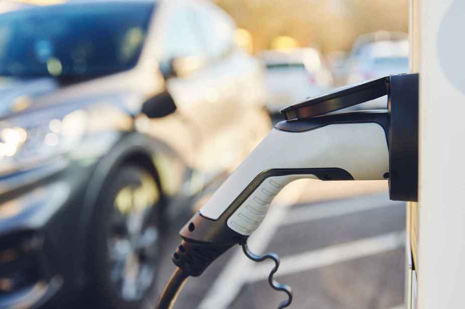 A white EV charger shown plugged into frame right while a blurred out car is parked with a charger plugged in the background