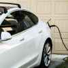 A white EV parked in front of a beige garage door at a residential home plugged into home EV charger