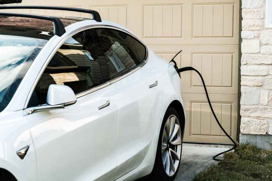 A white EV parked in front of a beige garage door at a residential home plugged into home EV charger