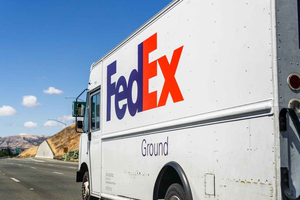A FedEx delivery truck driving on paved road left rear side angle view blue sky puffy clouds in background left of center