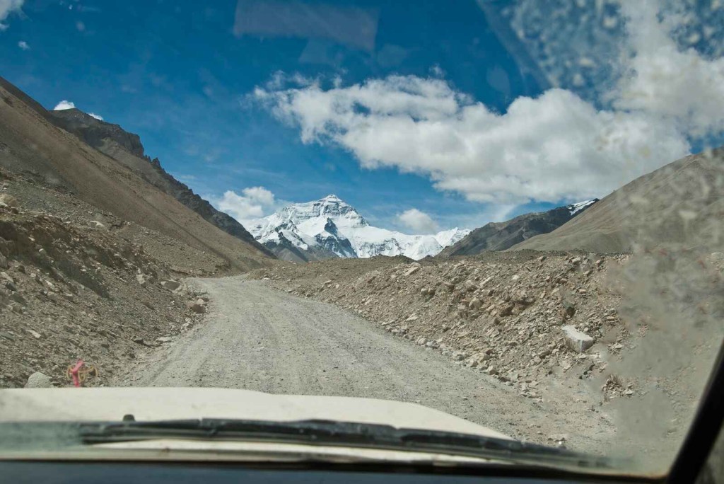 A view out of a white car windshield of a dirt road with Mount Everest in the midground blue cloudy sky