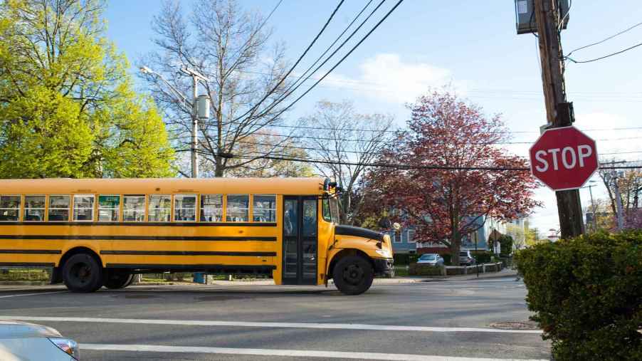 A school bus in nearly full right side profile view entering an intersection with a stop sign posted in the right midground and car nose peeking out of left rear corner