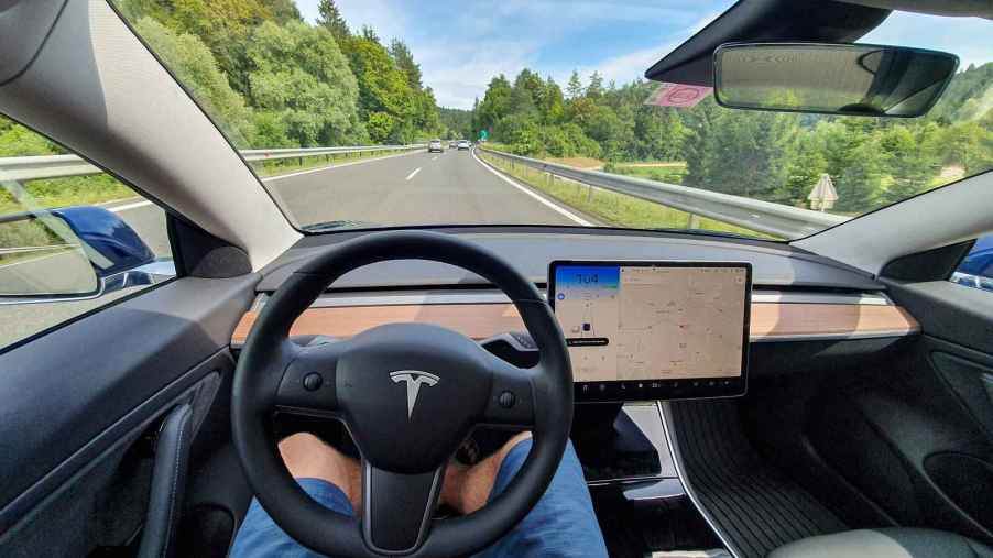 The driver interior view of a Tesla model in self driving mode man's legs under steering wheel no hands