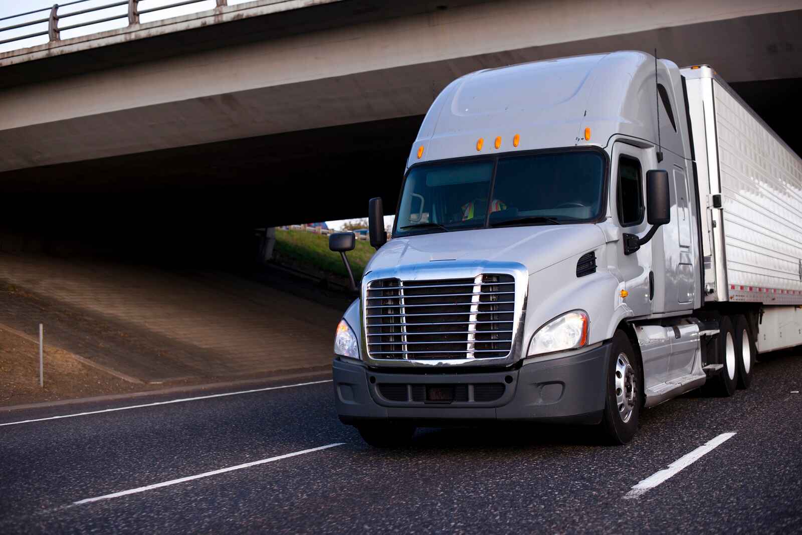 A white semi truck driving under an overpass bridge in close left front angle view
