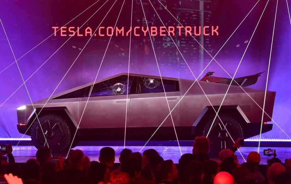A Tesla Cybertruck EV pickup is parked on a stage with crowd heads shown purple laser lighting and blue backlighting disco balls reflected in door glass