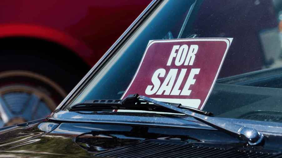 A red and white "FOR SALE" sign posted in a black car dash facing the windshield in close view.