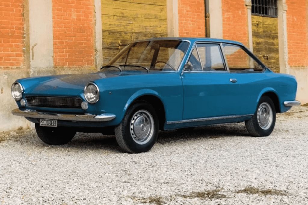 A blue 1967 Fiat 124 hardtop coupe parked in almost full left profile view parked on gravel